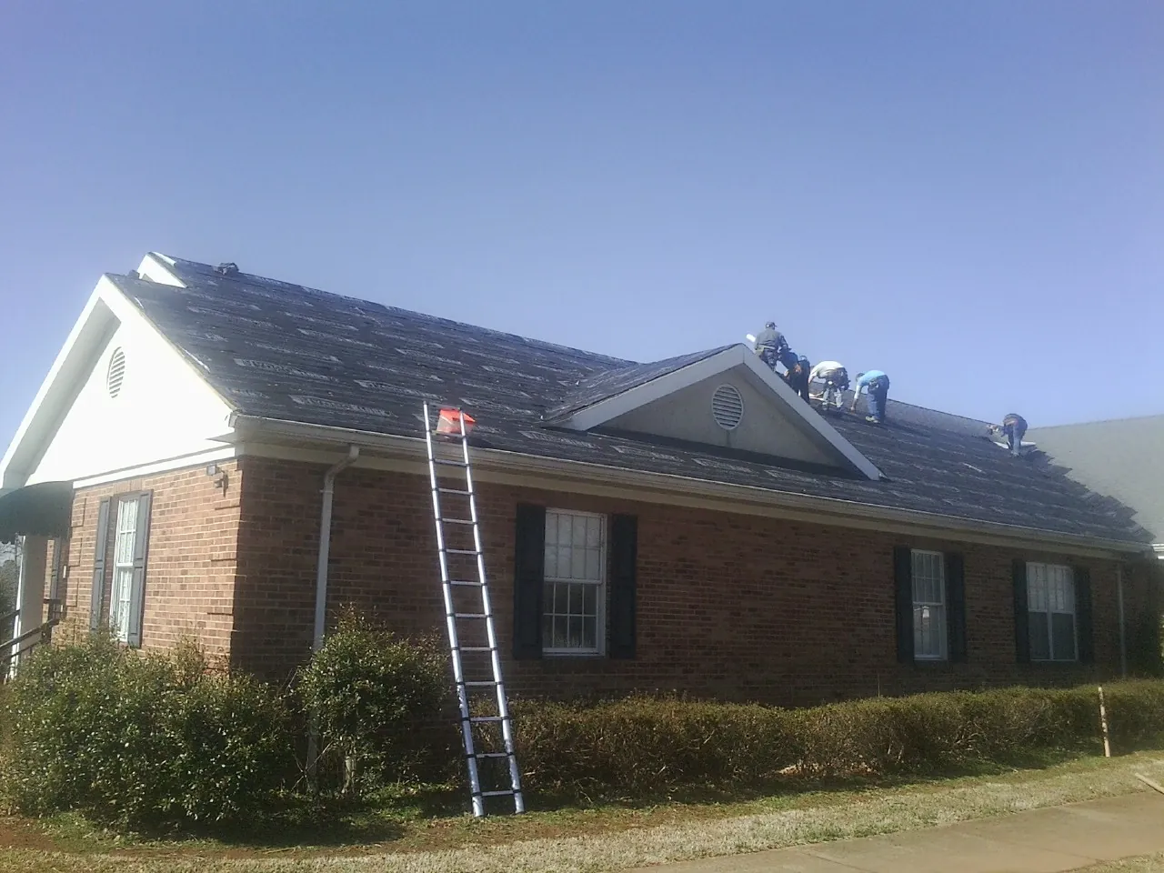 Crew tearing shingles off a brick ranch roof with a ladder set — Metal Roof Repair work by our crew in East Waterford