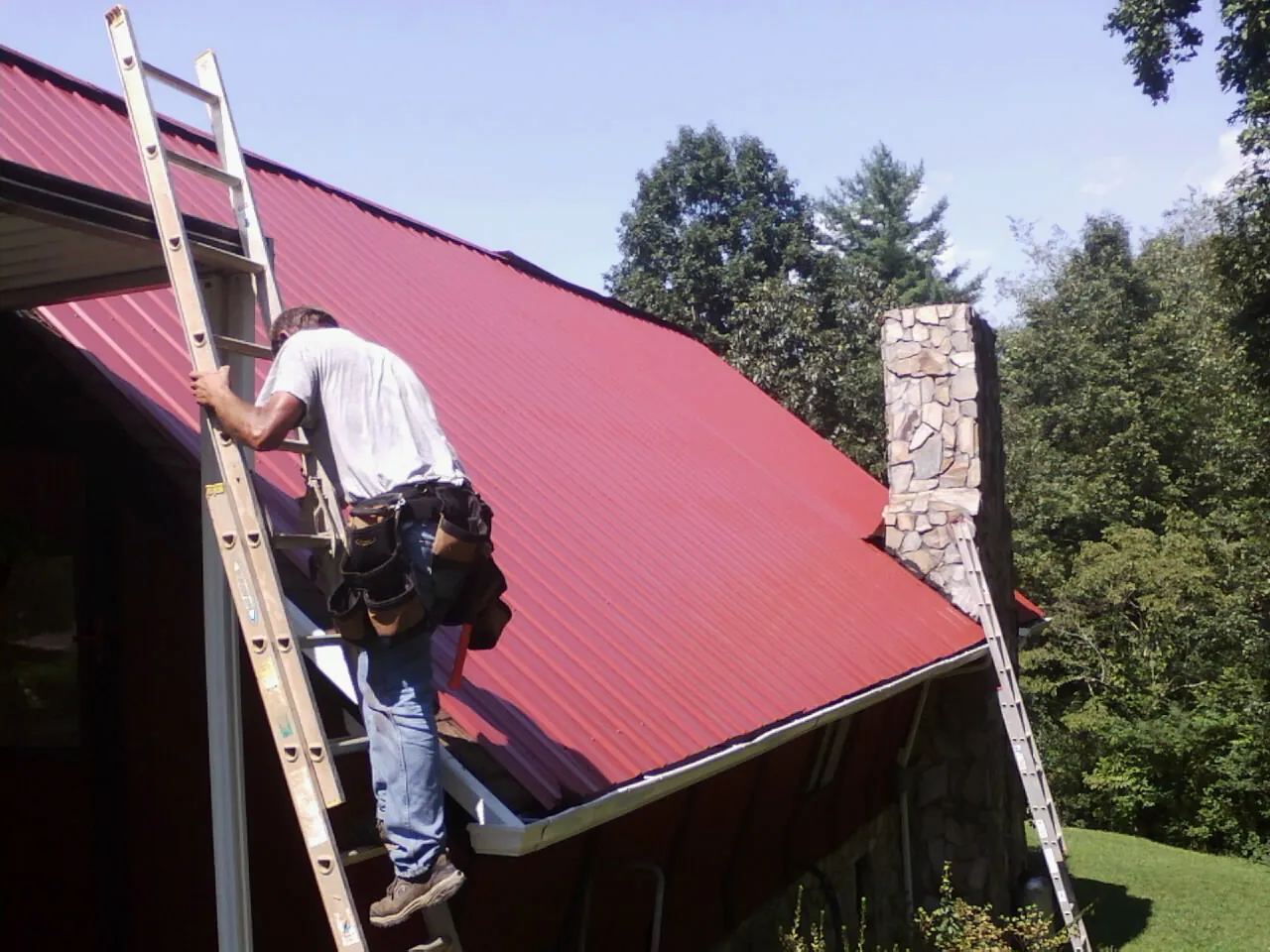 Roofer on a ladder beside a red metal roof and stone chimney — Soffit & Fascia Repair by Roofing Brothers in East Waterford